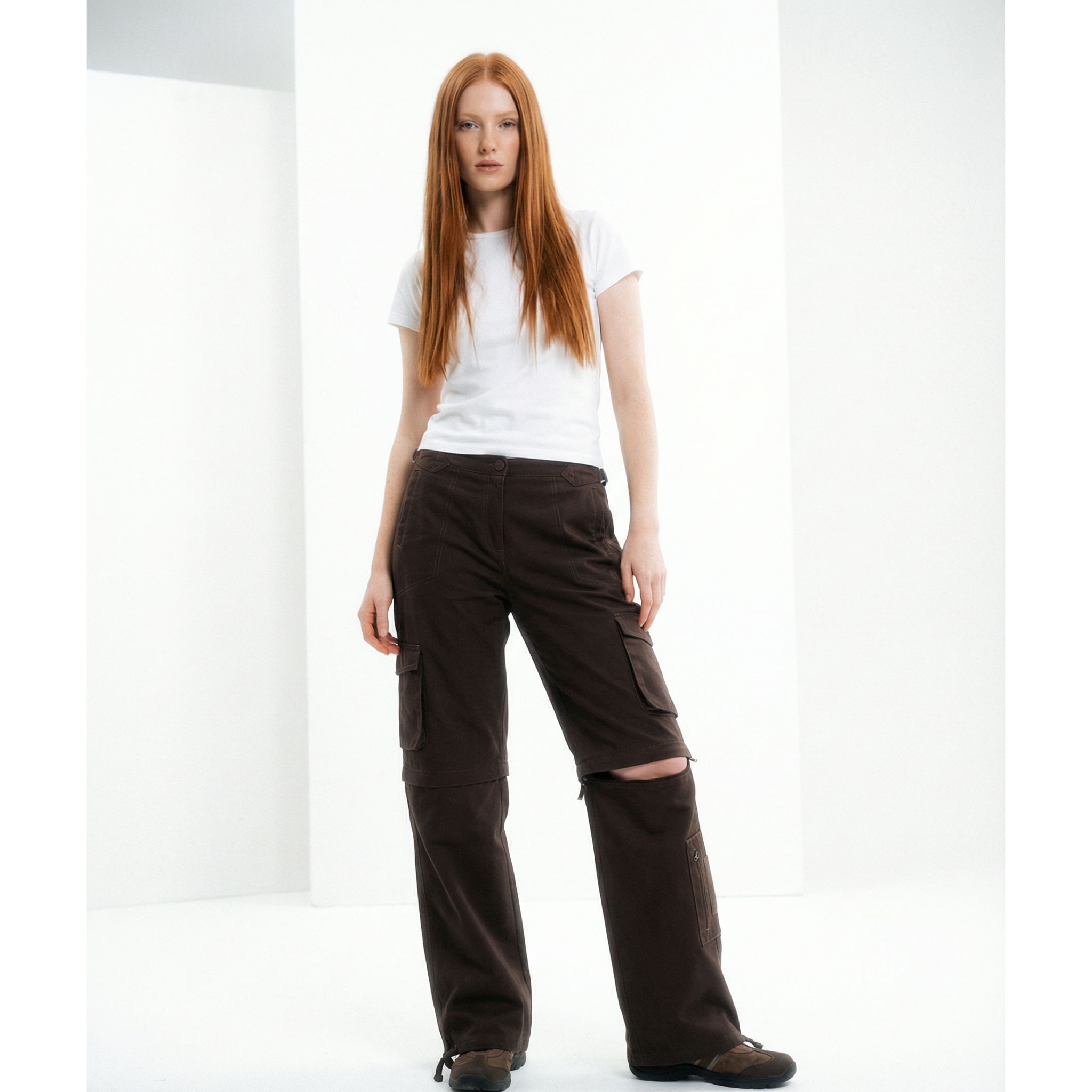 Red haired editorial model wearing brown utility cargo pants with a white tshirt. Convertible utility cargo pants zip-off detail is shown on one leg.